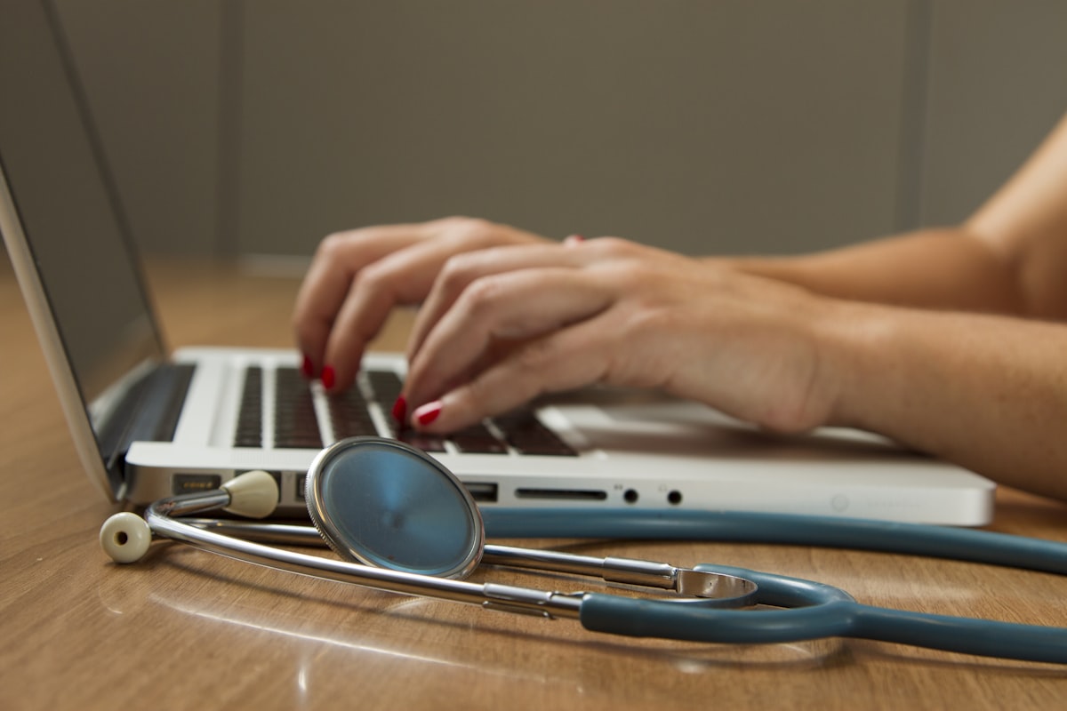 A clinician's hands on a laptop with a stethoscope beside it — a real provider on the other side of the screen.